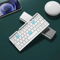 Silver white folding Bluetooth keyboard open next to a smartphone on its built-in metal stand.