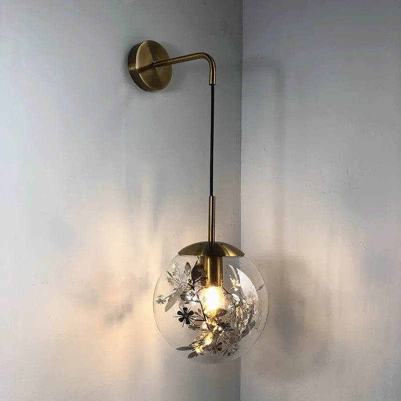 Modern minimalist glass wall lamp with golden flower finish, shown mounted beside a bed.