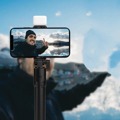 Black Bluetooth selfie stick with extended tripod legs, fill light, and a phone mounted in the 360-degree rotatable holder.