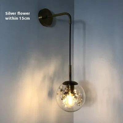 Modern minimalist glass wall lamp with golden flower finish, shown mounted beside a bed.