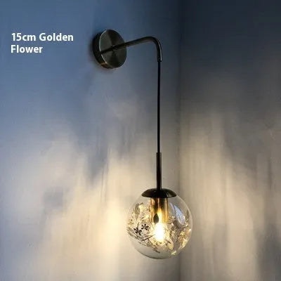 Modern minimalist glass wall lamp with golden flower finish, shown mounted beside a bed.