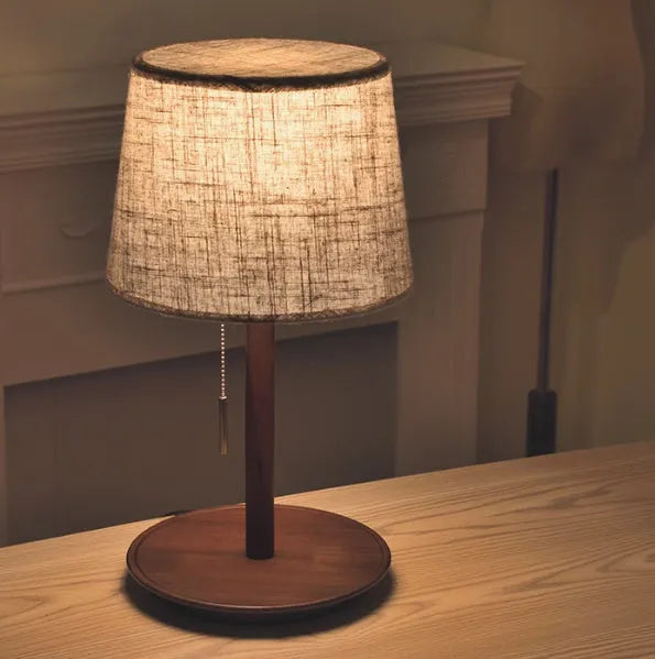 Walnut colour solid wood bedside lamp with white cloth shade on a wooden table.