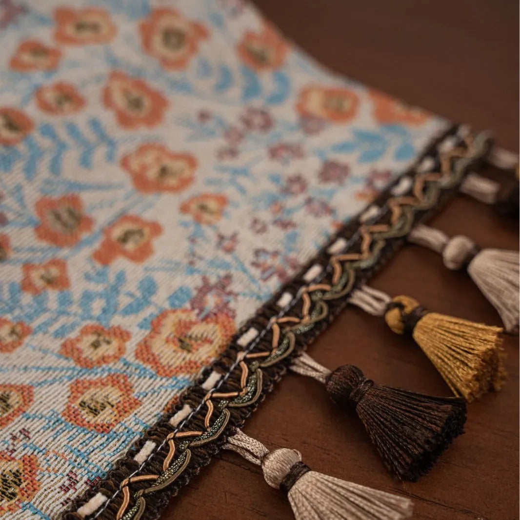 Beige jacquard table runner with floral pattern and long tassels on a wooden table.