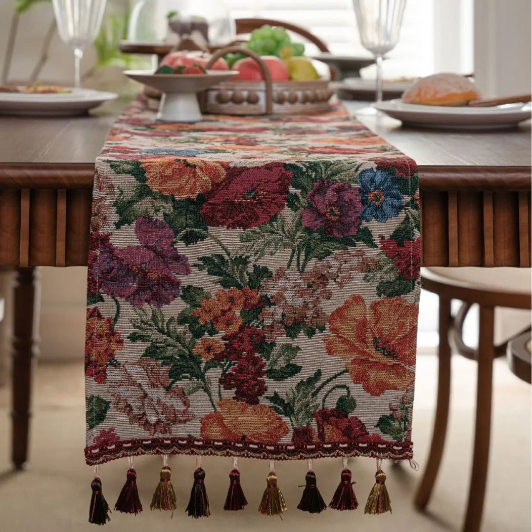 Beige jacquard table runner with floral pattern and long tassels on a wooden table.