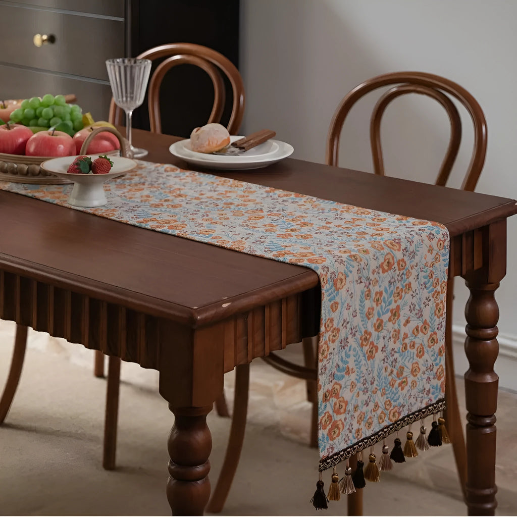Beige jacquard table runner with floral pattern and long tassels on a wooden table.