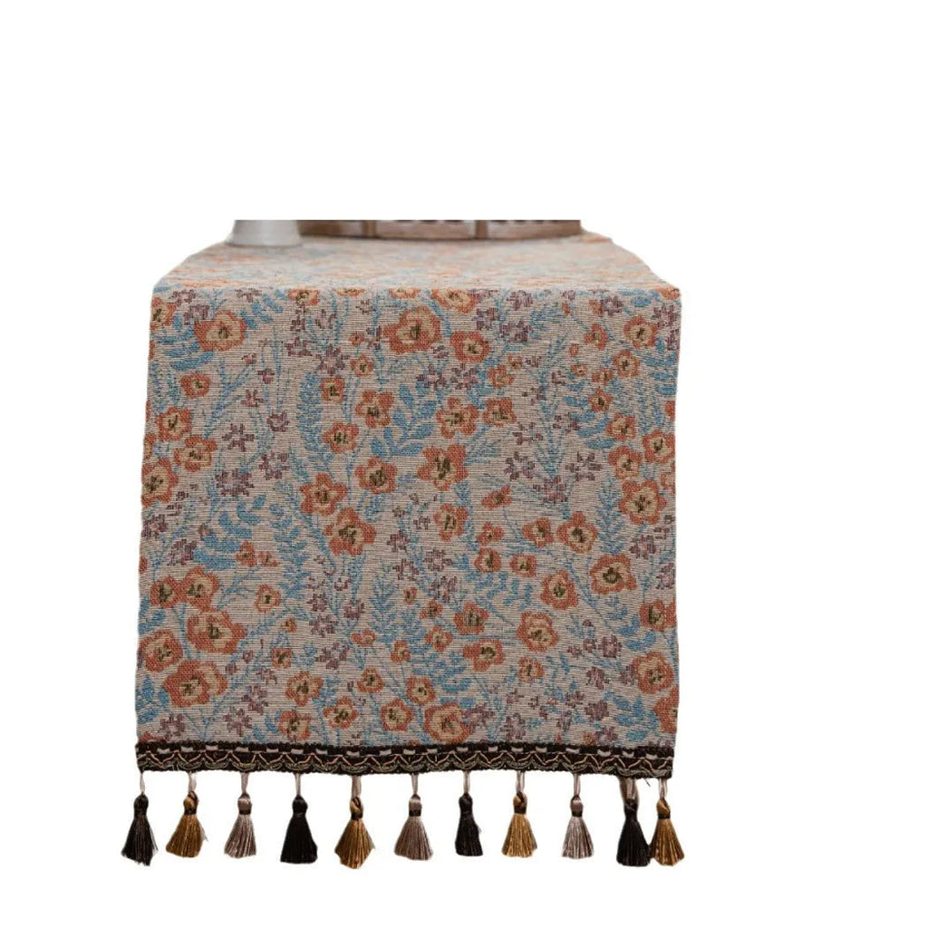 Beige jacquard table runner with floral pattern and long tassels on a wooden table.