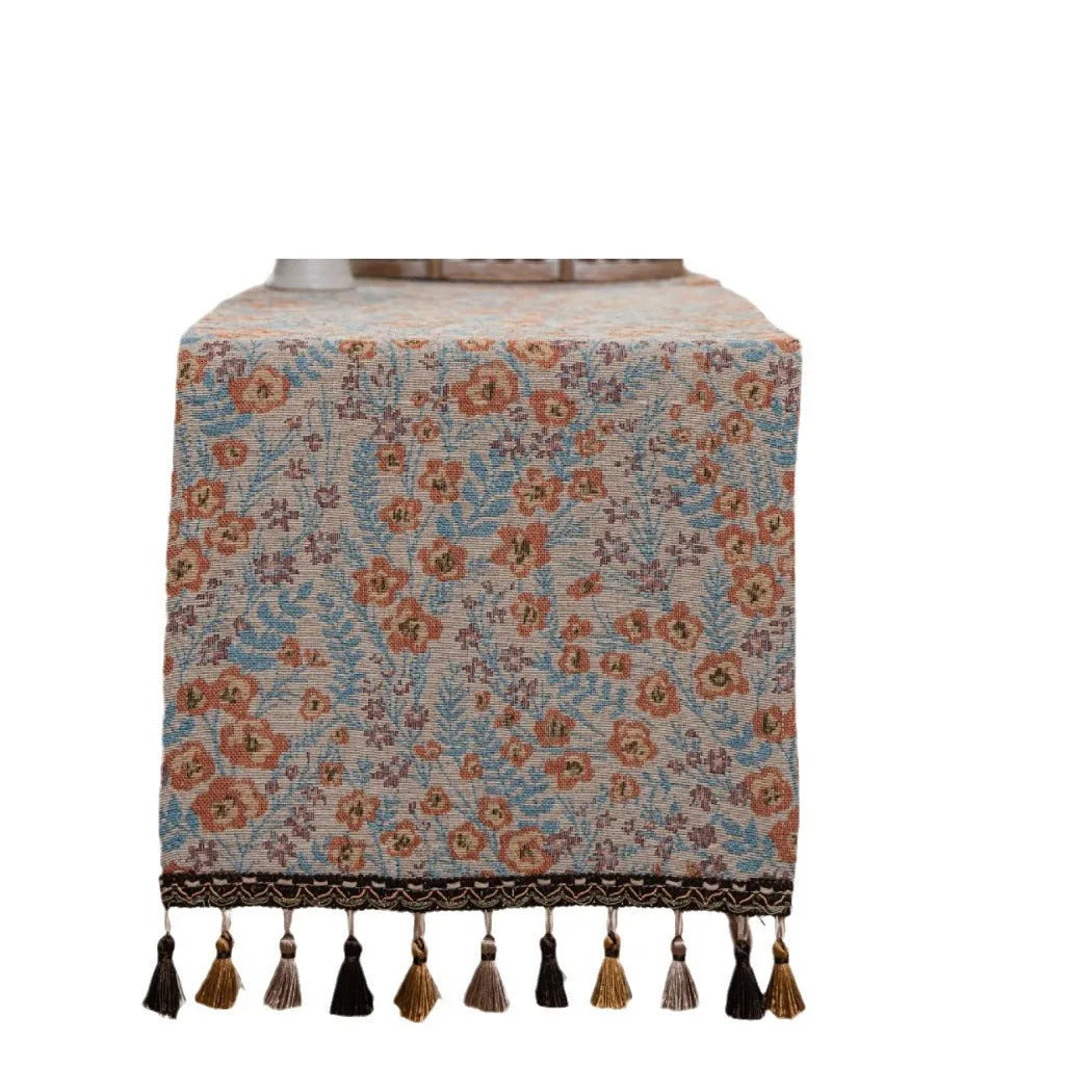 Beige jacquard table runner with floral pattern and long tassels on a wooden table.