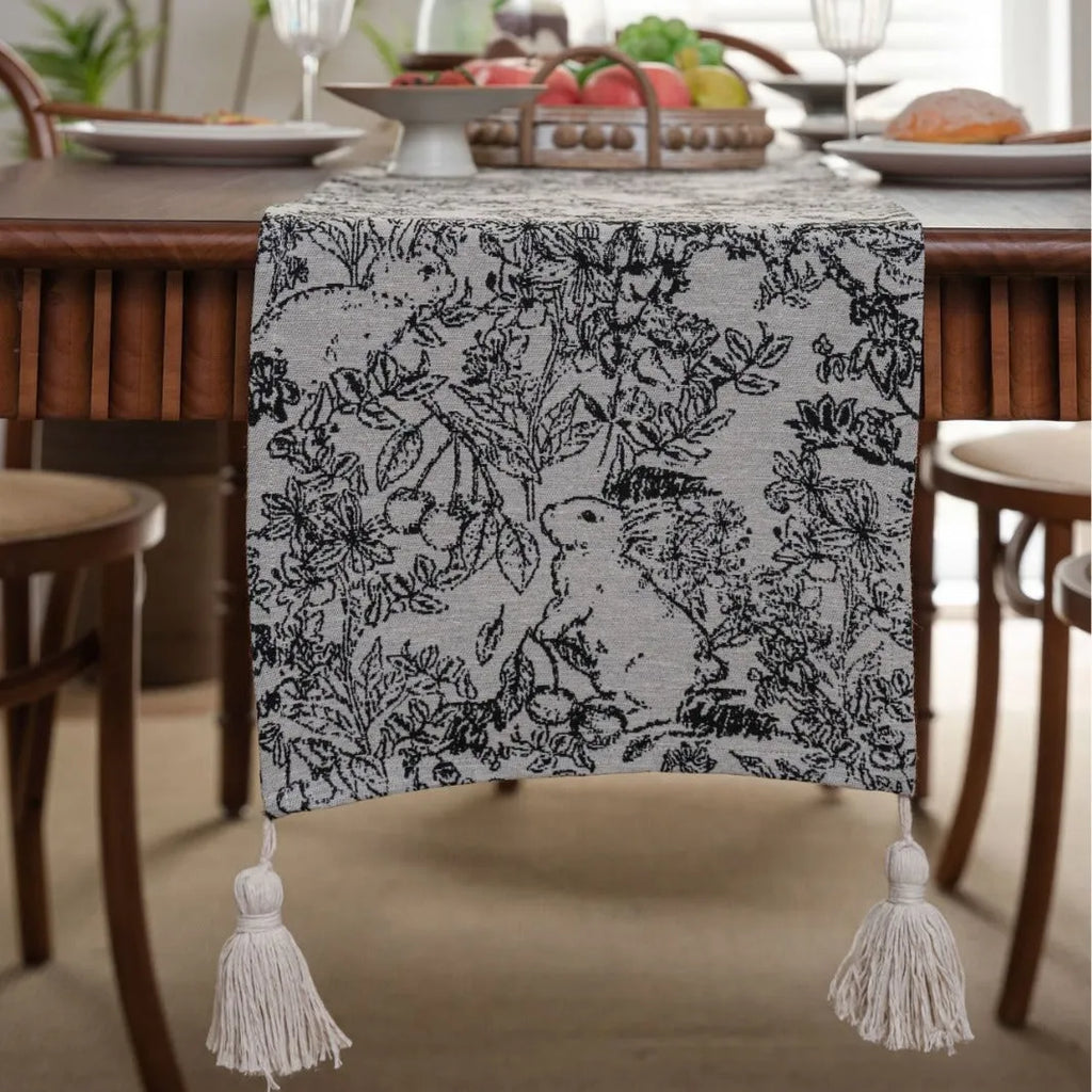 Beige jacquard table runner with floral pattern and long tassels on a wooden table.
