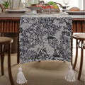 Beige jacquard table runner with floral pattern and long tassels on a wooden table.