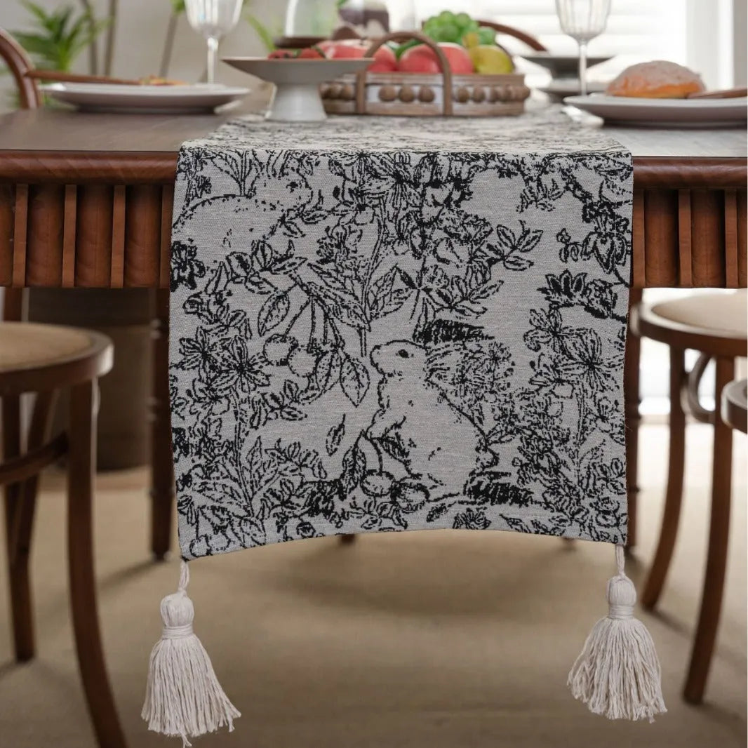 Beige jacquard table runner with floral pattern and long tassels on a wooden table.