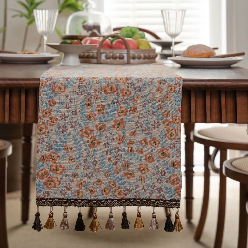 Beige jacquard table runner with floral pattern and long tassels on a wooden table.