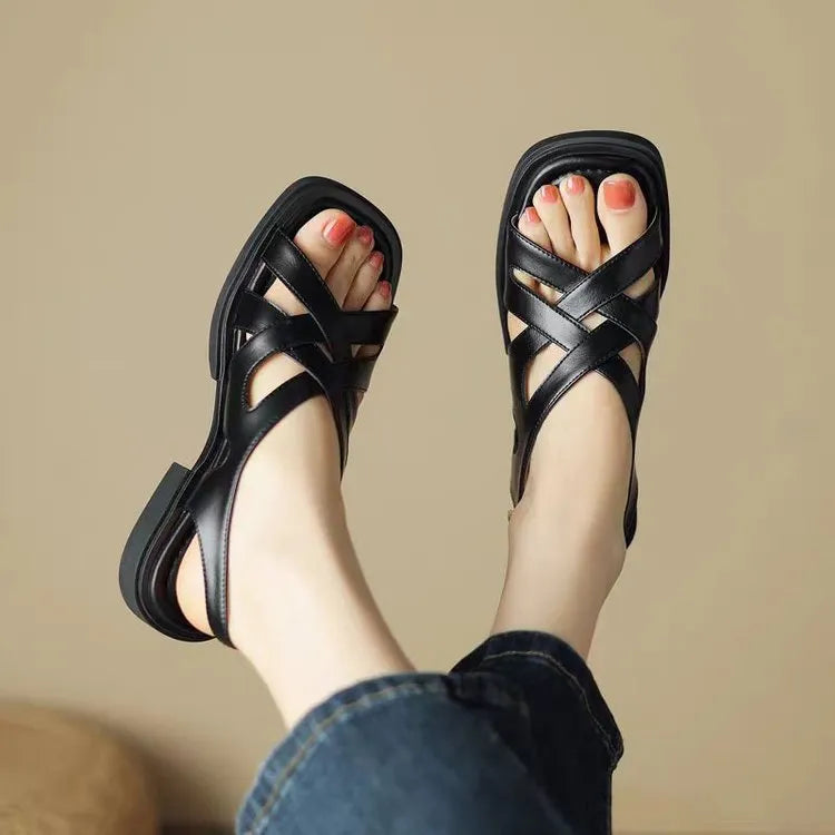Women's black patent leather square toe sandals with cross strap design on a beach background