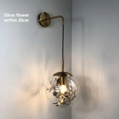 Modern minimalist glass wall lamp with golden flower finish, shown mounted beside a bed.