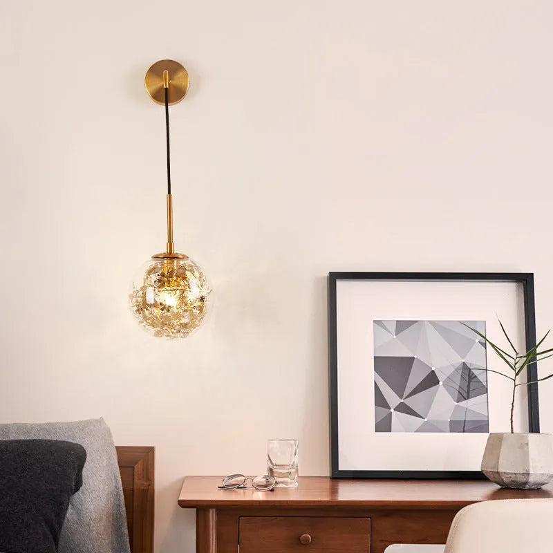 Modern minimalist glass wall lamp with golden flower finish, shown mounted beside a bed.