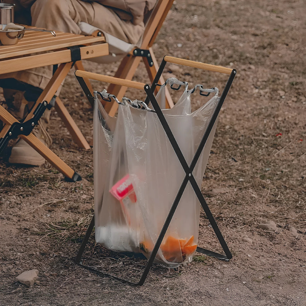 Folding black outdoor trash bag hanger with wooden carry handle on a patio.