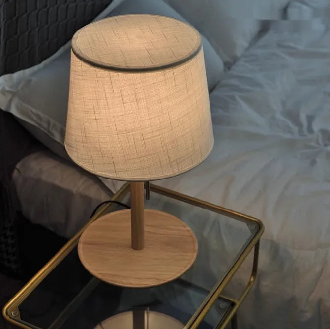 Walnut colour solid wood bedside lamp with white cloth shade on a wooden table.